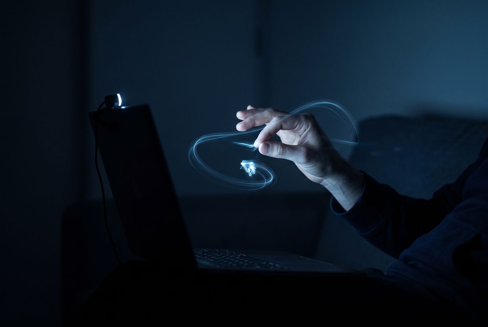 A hand in the air in front of a laptop, its fingertips leaving blue light trails